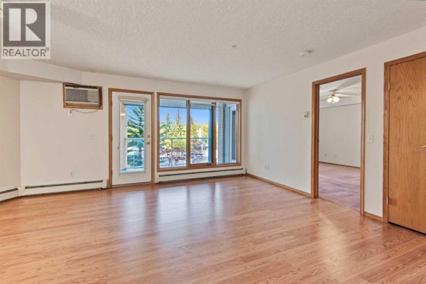 Living room with hardwood floors, glass door, and view of trees. - 220, 7229 Sierra Morena Boulevard Sw, Calgary