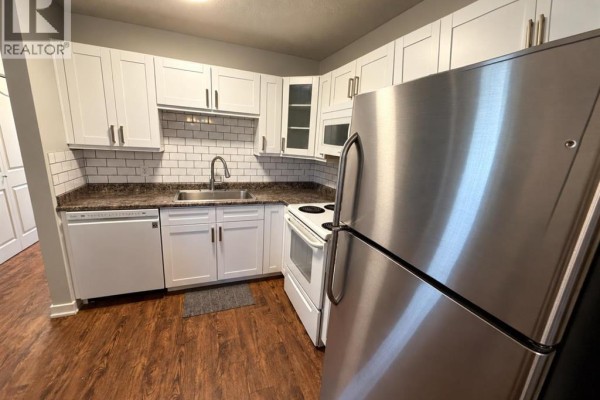 Compact kitchen with white cabinets, subway tile backsplash, stainless steel refrigerator, and dark wood floor. - 220 Mcgill Boulevard W, Lethbridge