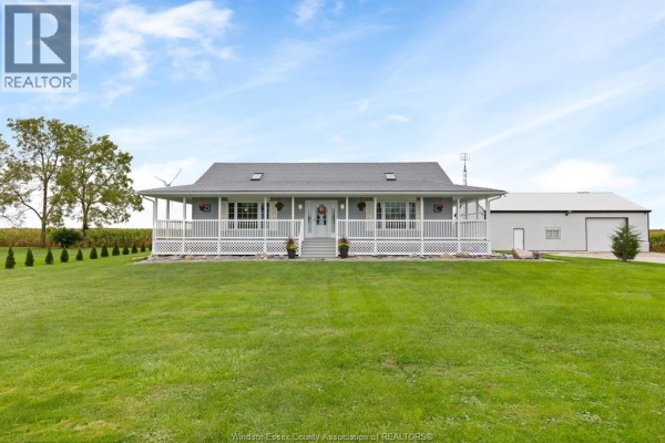 Front exterior of a light blue house with a white wrap-around porch, facing a wide green lawn and a large white garage. - 2208 County Rd 20 West, Essex