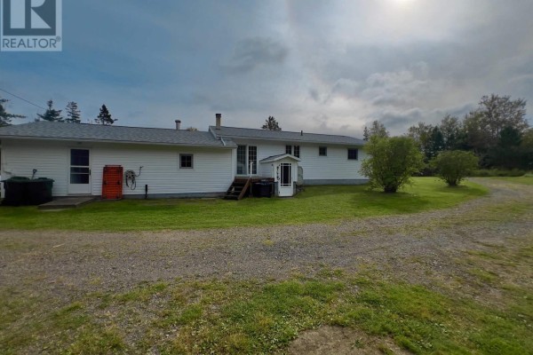 Rear exterior view of the white bungalow showing a gravel driveway and wooded area. - 2209 Lingan Road Road, Lingan