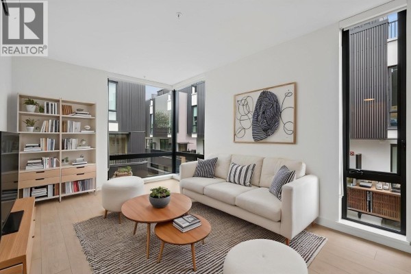 Living room with a sofa, coffee tables, bookshelf, and large windows overlooking the exterior. - 221 2820 Nanaimo Street, Vancouver