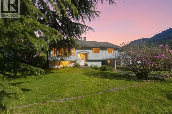 A light blue house with a driveway, grass lawn, and purple flowering bushes in the foreground. - 221 Spar St, Sayward