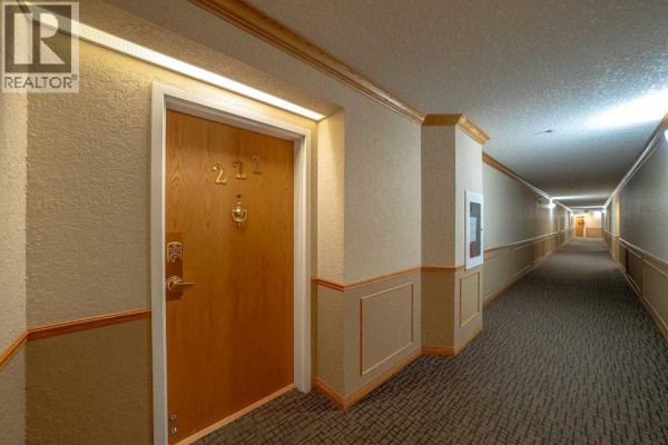 Apartment hallway with beige walls, patterned carpet, and a wooden door labeled 222. - 222, 6868 Sierra Morena Boulevard Sw, Calgary