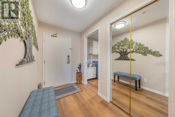 Apartment entryway with wood-look flooring, a blue bench, and mirrored closet doors. - 222, 6868 Sierra Morena Boulevard Sw, Calgary