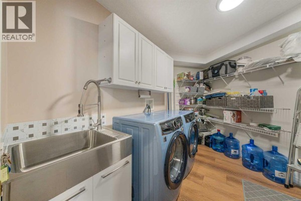 Laundry room with a large stainless steel sink, blue washer and dryer, and storage shelves. - 222, 6868 Sierra Morena Boulevard Sw, Calgary