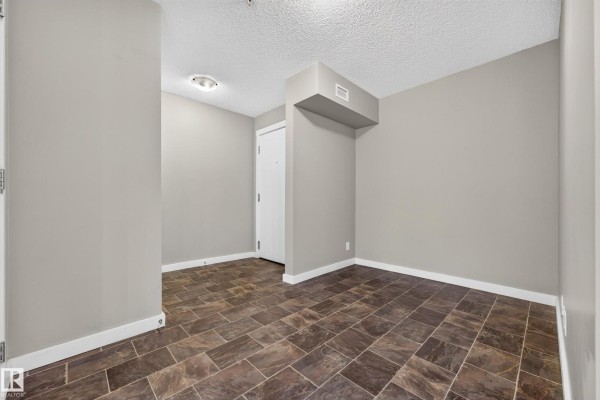 Interior view of an empty apartment room with beige walls and dark tile flooring. - #222 5810 Mullen Pl Nw, Edmonton