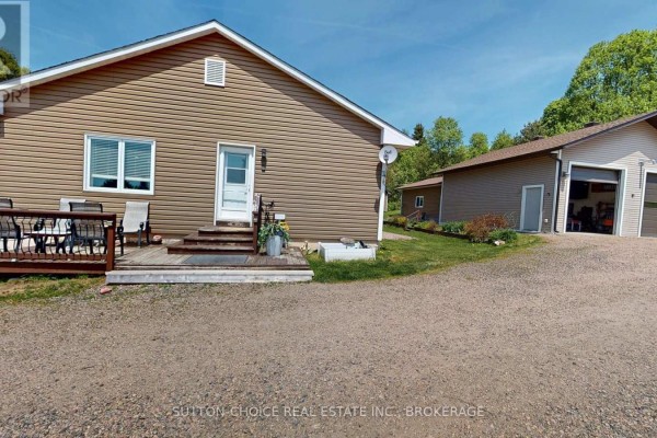 Side view of the beige-sided house and deck, with a gravel driveway and wooded background. - 222 Yonge Street, Bonfield