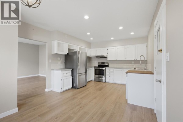 White kitchen with stainless steel appliances, light wood floors, and a doorway to another room. - 2231/2233 Valleyview Drive, Kamloops