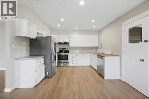 White kitchen with stainless steel appliances, light wood floors, and a doorway on the right. - 2231/2233 Valleyview Drive, Kamloops