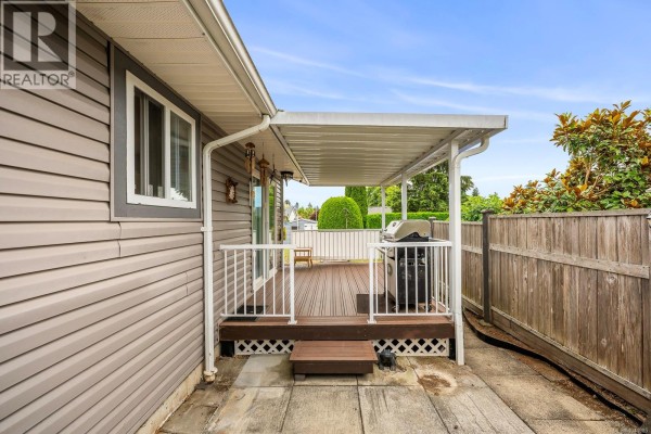 Covered deck with wooden planks and white railings next to a gray-sided house and a wooden fence. - 2233 6th St E, Courtenay
