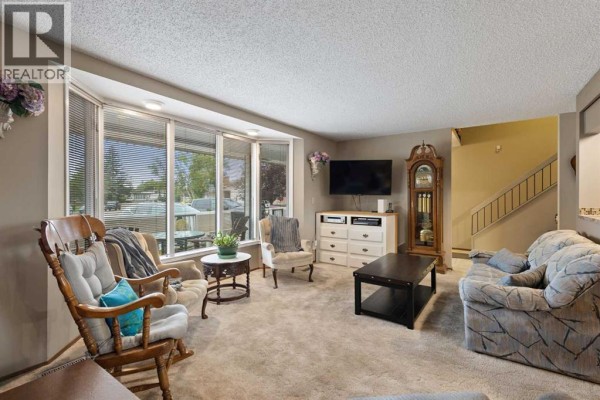 Living room with a large bay window, beige carpet, rocking chair, and grandfather clock. - 224 Pinetree Bay Ne, Calgary