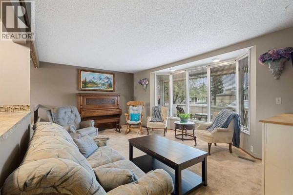 Living room with bay window, piano, multiple seating options, and beige carpet. - 224 Pinetree Bay Ne, Calgary