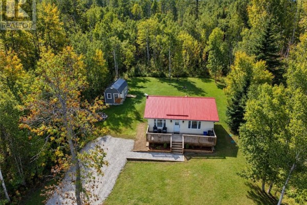 Aerial view of a white cottage with a red roof, wood deck, and a detached shed surrounded by a dense forest. - 2247 Bay Estates, Nemi