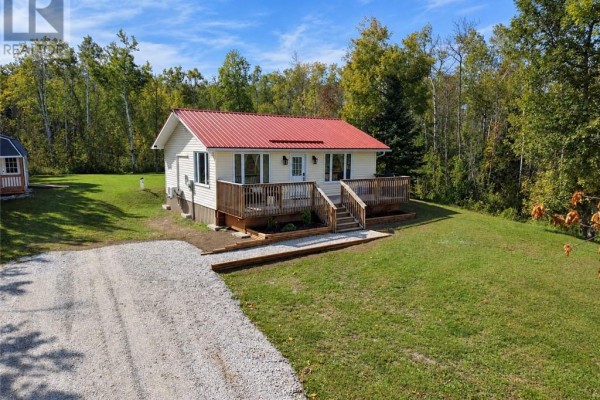 Front view of a white cottage with a red roof, wood deck, and a gravel driveway on a sunny day. - 2247 Bay Estates, Nemi