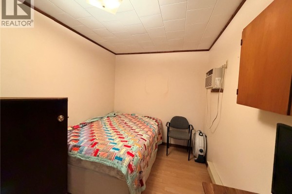 Bedroom with a quilt-covered bed, chair, small table, and wall-mounted air conditioner. - 225 5th Avenue W, Assiniboia