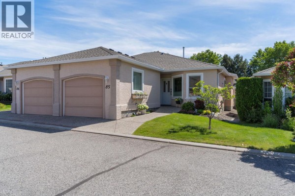 Exterior view of a beige stucco duplex with a double car garage and manicured lawn. - 2250 Louie Drive Unit# 85, Westbank