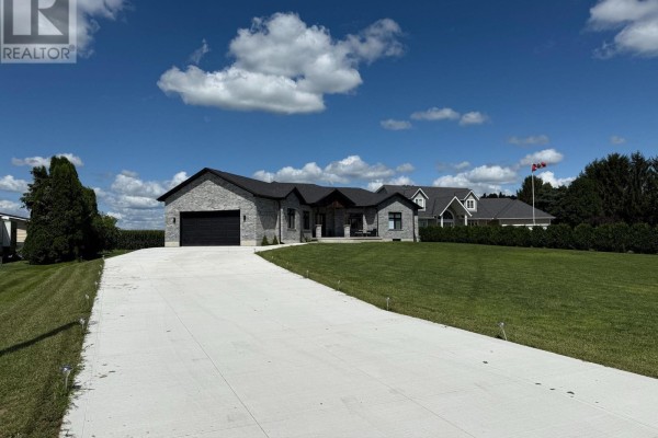 Long concrete driveway leading to a modern single-story house with a grey stone facade and attached garage. - 22664 Queens Line, West Elgin
