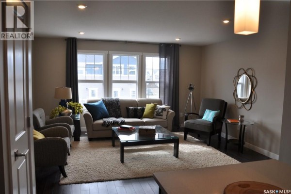 Living room with beige walls, large windows, gray sofa and chairs, coffee table, and rug. - 227 1220 Pringle Way, Saskatoon