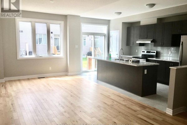 Spacious kitchen and living area with light wood floors, dark grey cabinets, stainless steel appliances, and an island. - 227 Bandelier Way, Ottawa