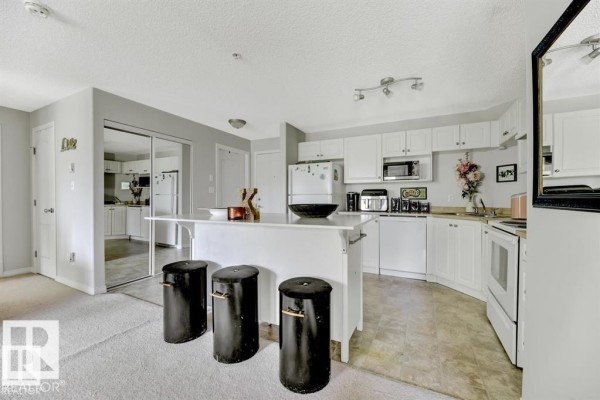 Open-concept apartment kitchen with white cabinets, island with bar stools, and stainless steel appliances. - #229 1520 Hammond Gate Nw, Edmonton