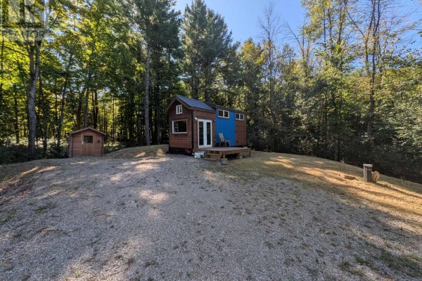 Front view of a small mobile home with blue siding and wooden exterior, set on a gravelly mound with trees in background. - 229 Norris Road, Tay Valley