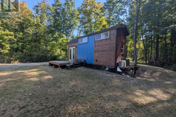 Rear three-quarter view of a mobile home with brown siding, wooden deck, and outdoor air conditioning unit, nestled in trees. - 229 Norris Road, Tay Valley