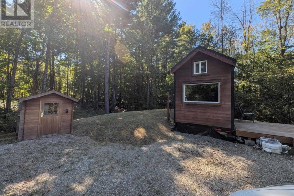 View of a small mobile home with a large window and wooden deck, next to a small wooden shed, with trees behind. - 229 Norris Road, Tay Valley