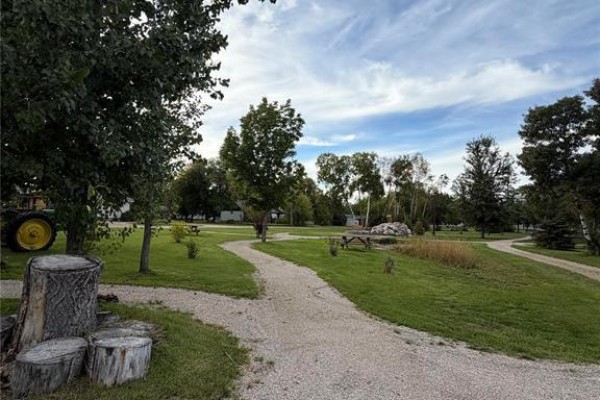 A gravel path curves through a park-like setting with trees, picnic tables, and a tractor on the left. - 23 1st Avenue S, Ashern