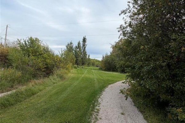 A gently curving gravel path passes through a green lawn and dense trees under a cloudy sky. - 23 1st Avenue S, Ashern
