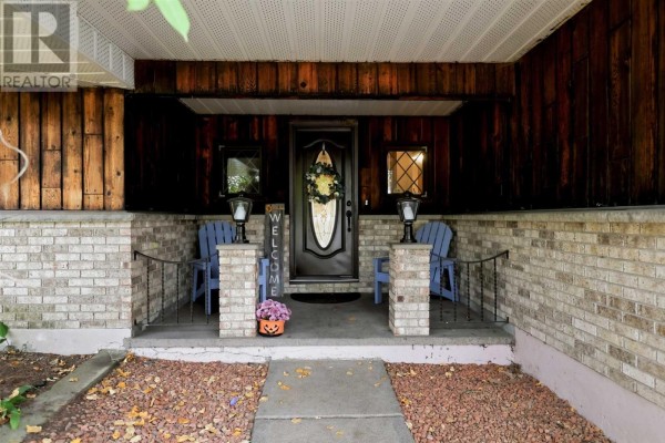 Covered front porch with double doors, brick pillars, two blue chairs, and glass lanterns. - 23 Country Club Pl, Sault Ste  Marie