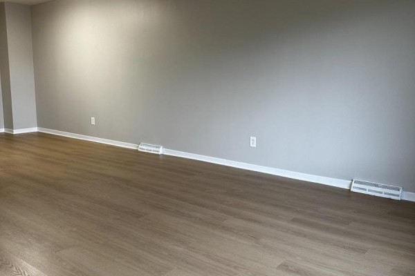 Empty room with light wood laminate flooring, neutral grey walls, and baseboard heating vents. - 23 Fairlawn Crescent, Welland