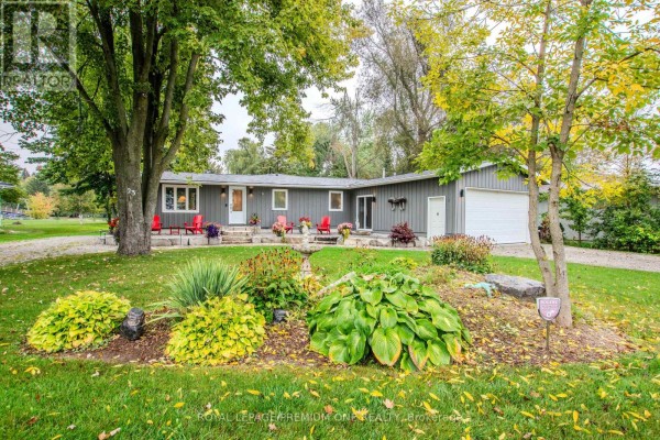 Exterior view of a grey bungalow with a prominent garden bed in the foreground and mature trees. - 23 Poplar Crescent, Ramara