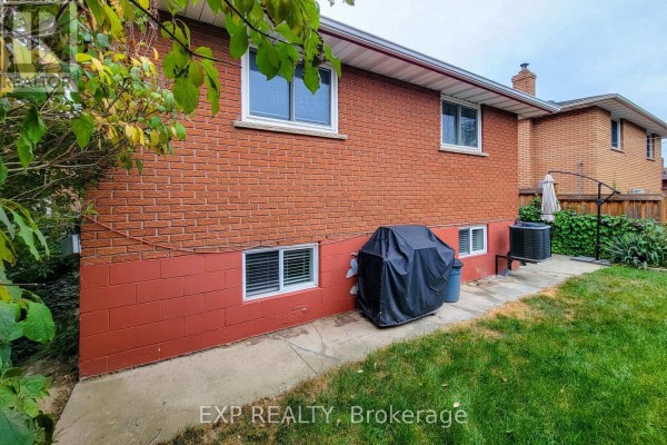 Close-up side view of the red brick house foundation and lower level windows, with outdoor appliances on the patio. - 23 Tara Court, Hamilton