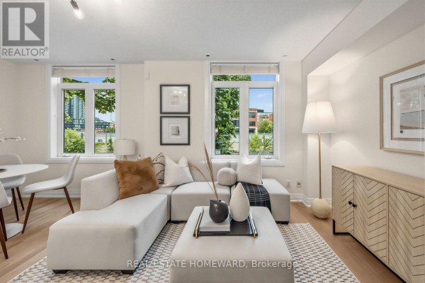 Living room with a white sectional sofa, coffee table, and large windows overlooking greenery. - 2306 - 23 Sudbury Street, Toronto