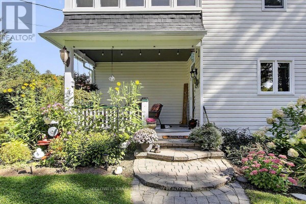 Close-up of the covered front porch with stone steps, potted plants, sunflowers, and a wicker chair visible inside. - 231 Boyds Road, Lanark Highlands