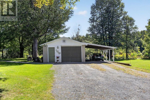 Detached garage with a double door and attached single carport, situated on a gravel driveway surrounded by grass and trees. - 231 Boyds Road, Lanark Highlands