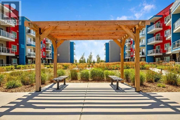 Outdoor common area with a wooden pergola, benches, and landscaped beds between apartment buildings. - 2313, 19489 Main Street Se, Calgary