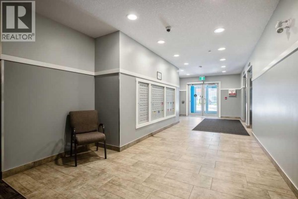 Interior building lobby with beige tiled floors, grey walls, a chair, mailboxes, and elevator doors. - 2313, 19489 Main Street Se, Calgary