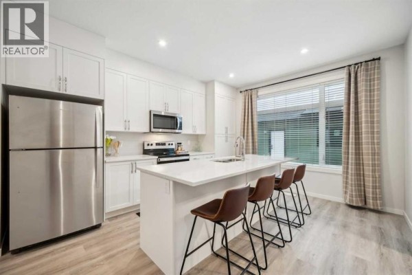 White kitchen with stainless steel appliances, island with bar stools, and checkered curtains. - 232 Vantage Drive, Cochrane