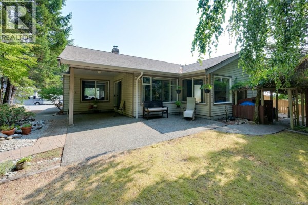 Backyard view of the beige house with a covered patio, lawn, and garden beds. - 2370 Larsen Rd, Shawnigan Lake