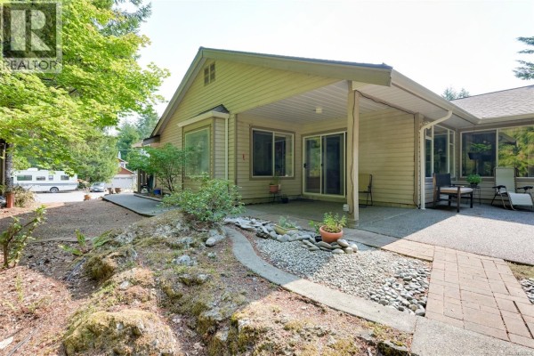 Side and backyard view of the house showing a covered patio, rock landscaping, and paved pathways. - 2370 Larsen Rd, Shawnigan Lake