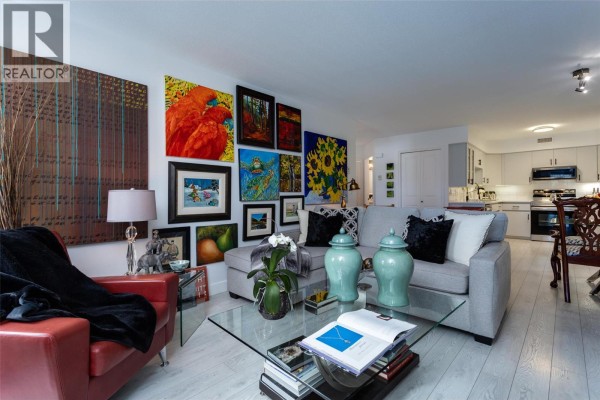 Living room with a sectional sofa, coffee table, fireplace, and kitchen in background. - 2350 Stillingfleet Road Unit# 113, Kelowna