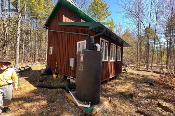 Man standing next to the cabin, showing the water tank and surrounding wooded area. - 2355 Raymo Road, Frontenac