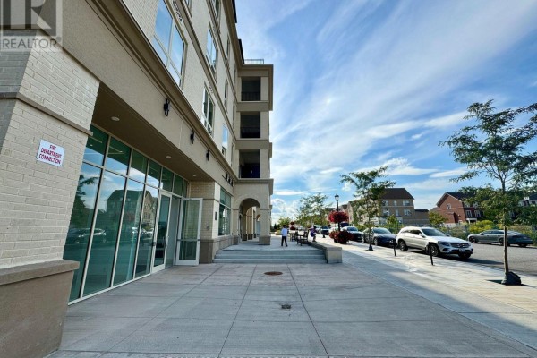 Close-up of a beige brick building facade with large glass windows and an arched entranceway on a sidewalk. - 239 - 101 Cathedral High Street, Markham