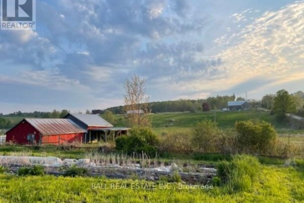 Red barn with a metal roof, surrounded by greenery and rolling hills under a cloudy sky. - 239 Concession 11w, Trent Hills
