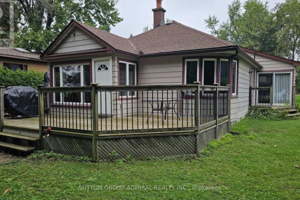 Rear exterior of a beige house with a wooden deck and dark railings. - 2392 Lakeshore Drive, Ramara