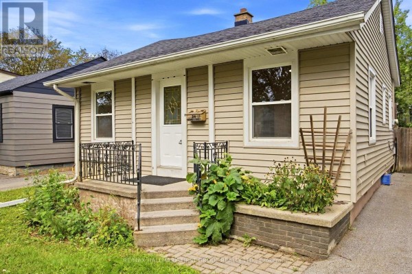 Front view of a beige bungalow with a small porch, steps, and surrounding greenery. - 24 George Street, Ajax