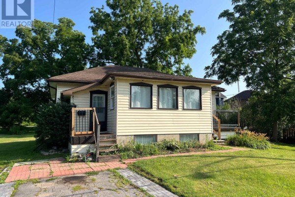 Rear view of a light yellow bungalow with dark trim, a small wooden deck, and surrounding grass. - 24 King Street W, Kawartha Lakes