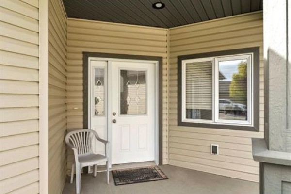 Front door of a beige house with sidelight windows and a white plastic chair. - 24 Wheatland Drive Se, Medicine Hat