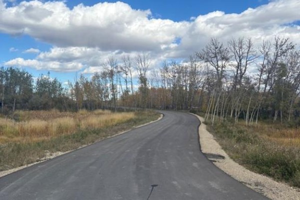 Paved road curves through a landscape with trees and dry grasses under a blue sky. - 24012 Aspen Drive, Rural Rocky View County